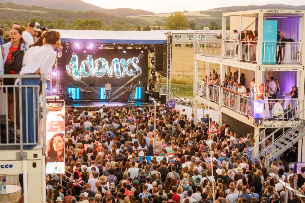 Koncert ADONXS na pódiu pri zóne IQOS Curious City na Pohode pri západe slnka, obrovský dav a ľudia na balkónoch kontajnerov