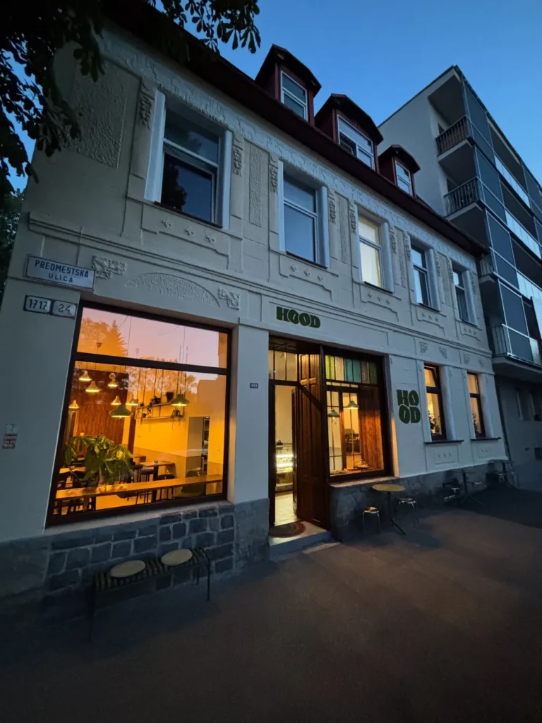 Rozsvietený interiér bistra HOOD v Žiline s logom na fasáde bývalého Art Café za súmraku